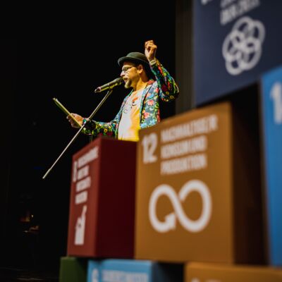 Bayerische Nachhaltigkeitstagung 2022 Nachhaltigkeit und Klimaschutz – notwendiger denn je!: Poetryslammer Michael Jakob steht auf der Bühne und fasst die Tagung in einem Poetry Slam zusammen